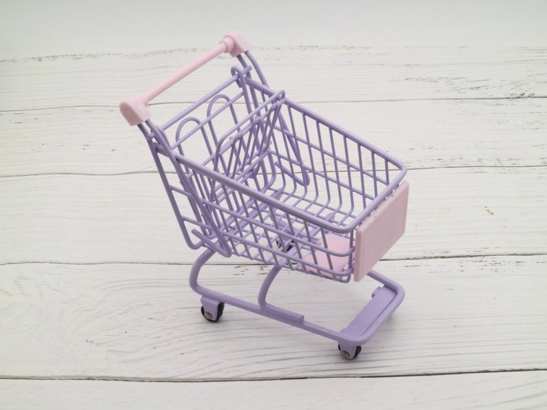 Puede incluir: Un peque&ntilde;o carro de la compra de color morado con un asa rosa y un panel frontal rosa. El carro est&aacute; sobre una superficie de madera blanca.