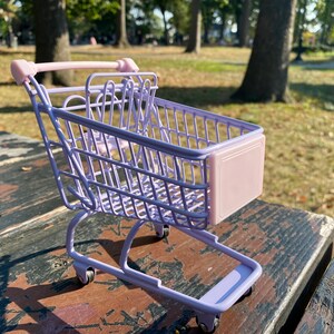 Puede incluir: Un peque&ntilde;o carro de la compra de color lavanda con un asa rosa y un panel frontal rosa. El carro est&aacute; sobre una superficie de madera.