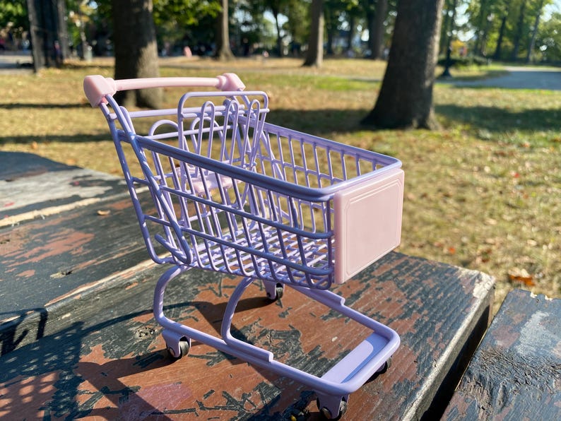 Puede incluir: Un peque&ntilde;o carro de la compra de color lavanda con un asa rosa y un panel frontal rosa. El carro est&aacute; sobre una superficie de madera.