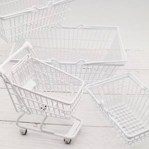 May include: Four white wire shopping baskets, one of which is a miniature shopping cart with wheels. The baskets are on a white wooden surface.