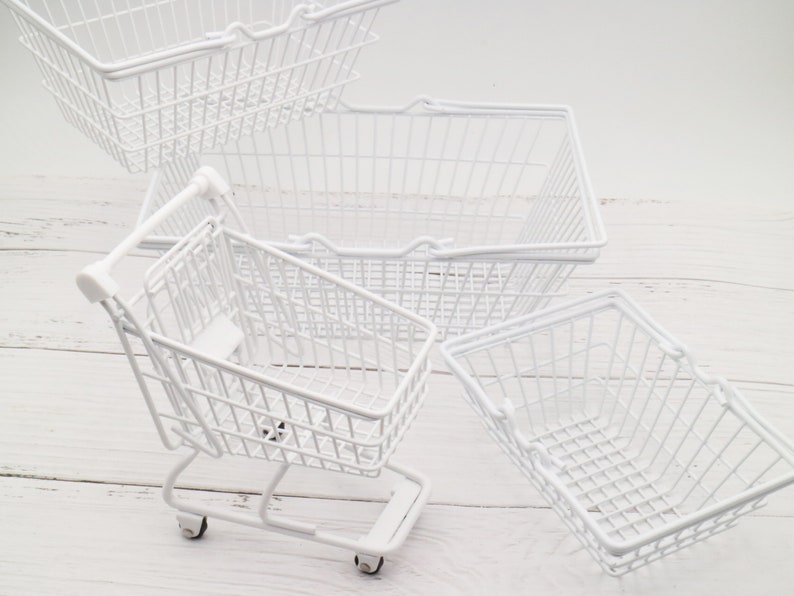 May include: Four white wire shopping baskets, one of which is a miniature shopping cart with wheels. The baskets are on a white wooden surface.