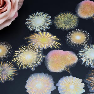 Peut inclure: Une collection d'autocollants transparents représentant divers motifs de feux d'artifice aux tons or, jaune et rose. Une grande rose rose clair est dans le coin supérieur gauche, sur fond sombre.