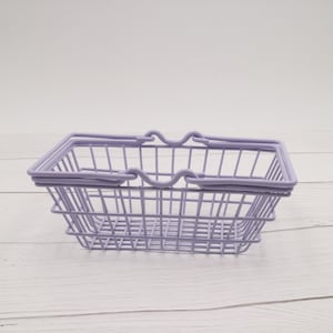 Puede incluir: Una cesta de la compra de alambre lavanda con dos asas. La cesta est&aacute; vac&iacute;a y se encuentra sobre una superficie de madera blanca.