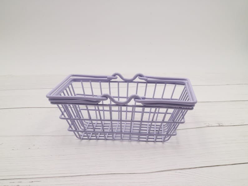 Puede incluir: Una cesta de la compra de alambre lavanda con dos asas. La cesta est&aacute; vac&iacute;a y se encuentra sobre una superficie de madera blanca.