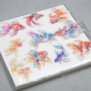 May include: A collection of colorful goldfish stickers on a white notebook. The stickers feature a variety of vibrant colors, including red, orange, blue, and yellow. The notebook has a clear plastic cover and a silver metal binding.