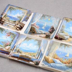 May include: A collection of six square art prints depicting serene coastal scenes viewed through open windows. Each print showcases a vibrant blue sky, ocean, and various outdoor settings with furniture and floral arrangements. The prints are arranged in an open book.