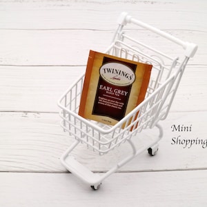 May include: A white miniature shopping cart with a single tea bag inside. The tea bag is for "Twinings Earl Grey Black Tea". The cart is on a white wooden surface.
