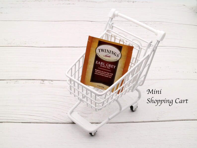 May include: A white miniature shopping cart with a single tea bag inside. The tea bag is for "Twinings Earl Grey Black Tea". The cart is on a white wooden surface.