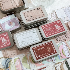 Może przedstawiać: Kilka małych, prostokątnych metalowych puszek z ozdobnymi etykietami, niektóre ułożone w stos. Etykiety zawierają tekst i ilustracje w odcieniach czerwieni, bieli i brązu. Różne papierowe efemerydy o podobnych wzorach otaczają puszki.