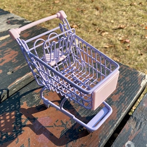 Puede incluir: Un peque&ntilde;o carrito de la compra de color lavanda con un asa rosa y una base rosa. El carrito est&aacute; hecho de alambre met&aacute;lico y tiene una forma rectangular.