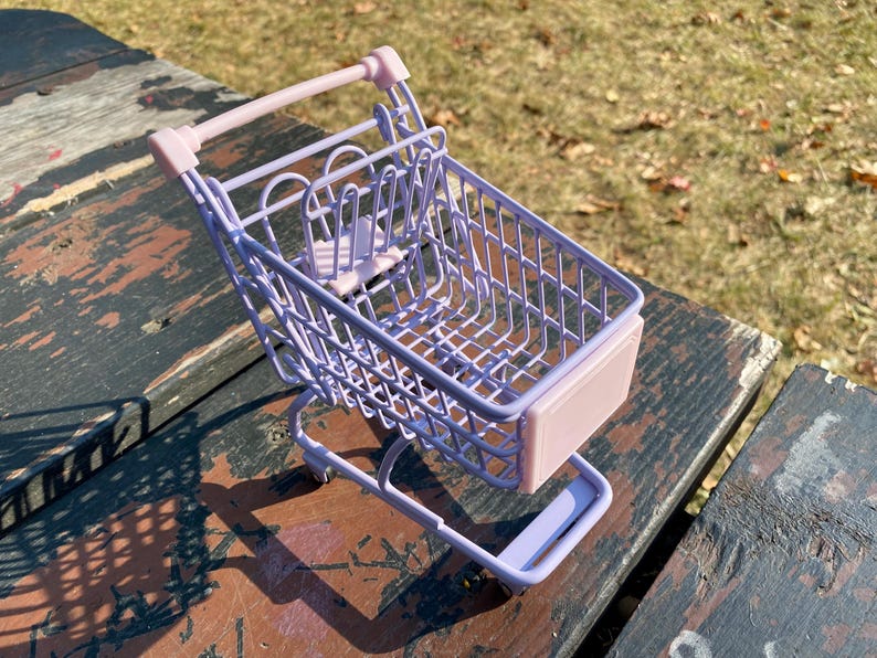 Puede incluir: Un peque&ntilde;o carrito de la compra de color lavanda con un asa rosa y una base rosa. El carrito est&aacute; hecho de alambre met&aacute;lico y tiene una forma rectangular.