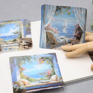 May include: Three square stickers with scenic paintings of coastal views. One shows a piano and chair by a window, another a sailboat on the water, and the third a window seat with pillows and a book. The stickers are on a white surface.