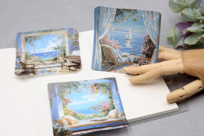 May include: Three square stickers with scenic paintings of coastal views. One shows a piano and chair by a window, another a sailboat on the water, and the third a window seat with pillows and a book. The stickers are on a white surface.