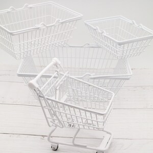 May include: Four white wire shopping baskets, one with wheels, stacked on a white wooden surface. The baskets are empty.