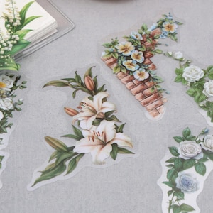 May include: A collection of transparent decorative stickers featuring botanical illustrations of white lilies, roses, daisies, and lily of the valley, along with a brick wall motif, arranged in various floral clusters and trailing vine patterns.