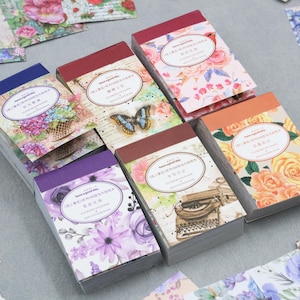 May include: An assortment of floral-themed stationery sets. Each set features a different floral design, including roses, hydrangeas, and butterflies. The sets have a rectangular shape and are stacked together. The text "Have a good day" is printed on the top of each set.