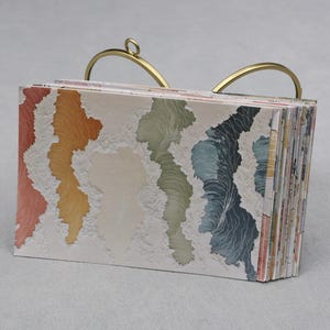 May include: A small, colorful notebook with a gold-toned handle. The pages display a watercolor-like design in shades of red, orange, yellow, green, and blue. The notebook is open, revealing the patterned pages.