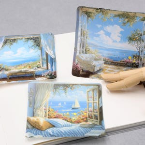 May include: Three rectangular art pieces depicting scenic coastal views. Each piece showcases a window or open space with a blue sky, ocean, and lush greenery. One features a sailboat, another a patio with a white couch, and the third a table.