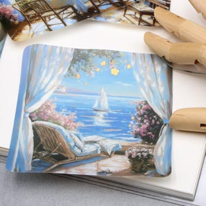 May include: A small, rectangular print depicts a serene seaside scene. A sailboat glides on the blue water, framed by white curtains and pink flowers. A wicker chair and a basket of flowers are also visible.
