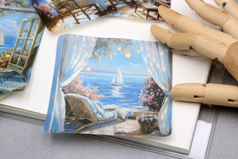 May include: A small, rectangular print depicts a serene seaside scene. A sailboat glides on the blue water, framed by white curtains and pink flowers. A wicker chair and a basket of flowers are also visible.