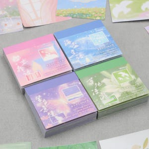 May include: Four stacks of pastel-colored sticky notes with dreamy illustrations. Each stack has a different color: pink, blue, green, and purple. The top notes feature images of landscapes and text. Other loose notes with similar designs are scattered around.