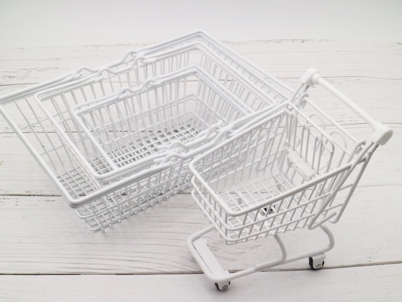 May include: Four white wire shopping baskets, one of which is a miniature shopping cart with wheels. The baskets are stacked on top of each other.