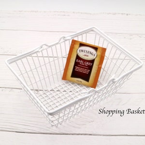 May include: White wire shopping basket with a single tea bag inside. The tea bag is labeled "Twinings Earl Grey Black Tea". The text "Shopping Basket M" is written below the basket.