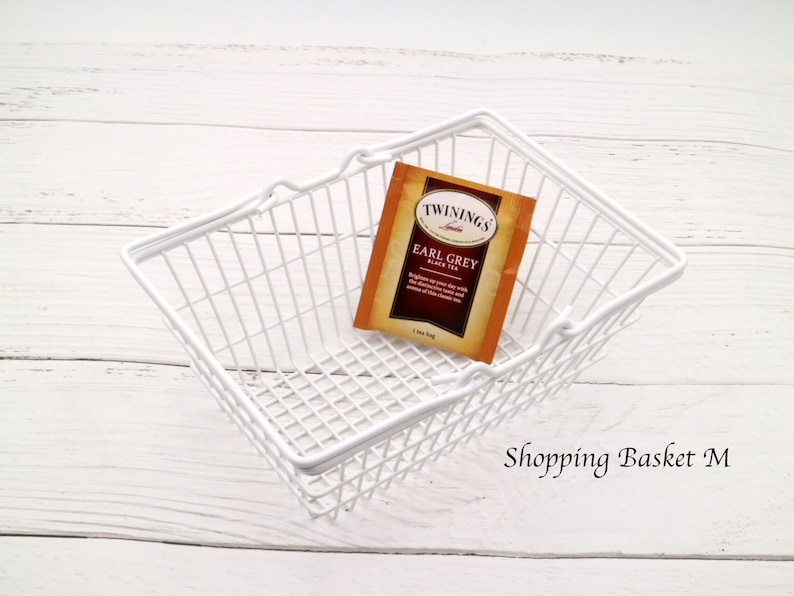 May include: White wire shopping basket with a single tea bag inside. The tea bag is labeled "Twinings Earl Grey Black Tea". The text "Shopping Basket M" is written below the basket.