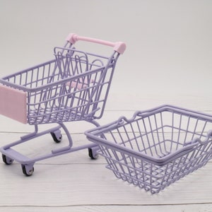 Puede incluir: Dos carros de la compra de metal morado. Uno es un carro de la compra en miniatura con ruedas y un asa rosa. El otro es una cesta m&aacute;s peque&ntilde;a con un asa.