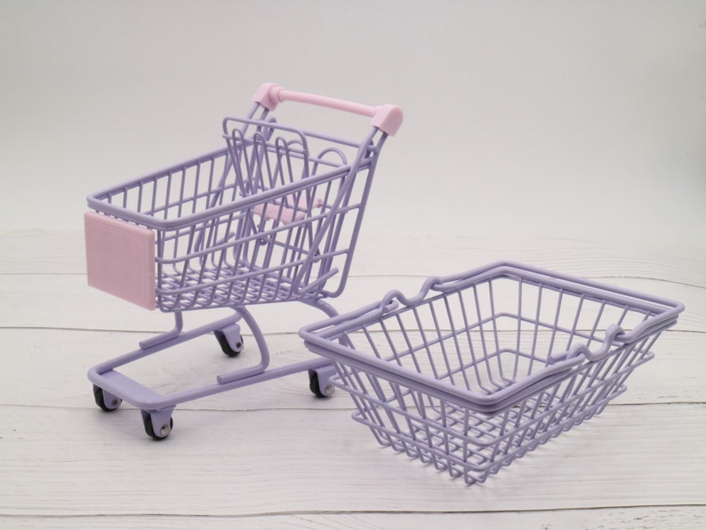 Puede incluir: Dos carros de la compra de metal morado. Uno es un carro de la compra en miniatura con ruedas y un asa rosa. El otro es una cesta m&aacute;s peque&ntilde;a con un asa.