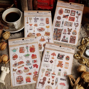 以下が含まれることがあります： 中世風の様々なオブジェクトを描いたステッカーが4枚セットになっています。ステッカーはベージュの縁取りのある白い紙に印刷されており、上部に「Medieval Collection Room」の文字が印刷されています。ステッカーには、ティーカップ、ティーポット、ケーキ、ペストリー、本、キャンドルなどのオブジェクトのイラストが描かれています。