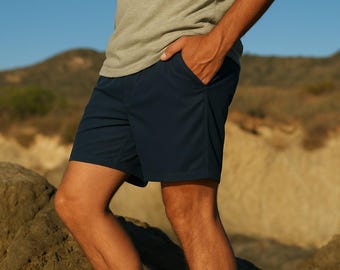 Puede incluir: Una persona lleva un par de pantalones cortos deportivos azul oscuro y una camiseta verde claro. Lleva un par de zapatillas de lona verde oscuro con cordones blancos. La persona está de pie sobre una superficie rocosa.