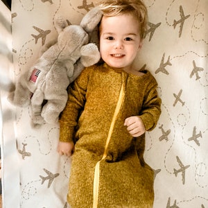 May include: A young child wearing a mustard yellow sleep sack with a yellow zipper is lying in a cot. The cot sheets have a pattern of grey aeroplanes on a white background. The child is holding a grey stuffed elephant with a small American flag on its side.