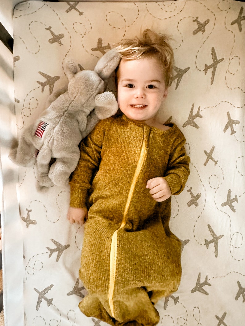 May include: A young child wearing a mustard yellow sleep sack with a yellow zipper is lying in a cot. The cot sheets have a pattern of grey aeroplanes on a white background. The child is holding a grey stuffed elephant with a small American flag on its side.
