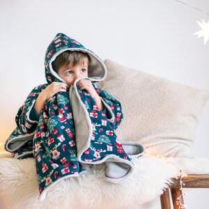 May include: A child wearing a hooded, blue and white fleece blanket with a Christmas print. The blanket has a red, white, and green pattern with images of Christmas trees, presents, and reindeer. The child is sitting on a white furry surface with a beige pillow behind them.