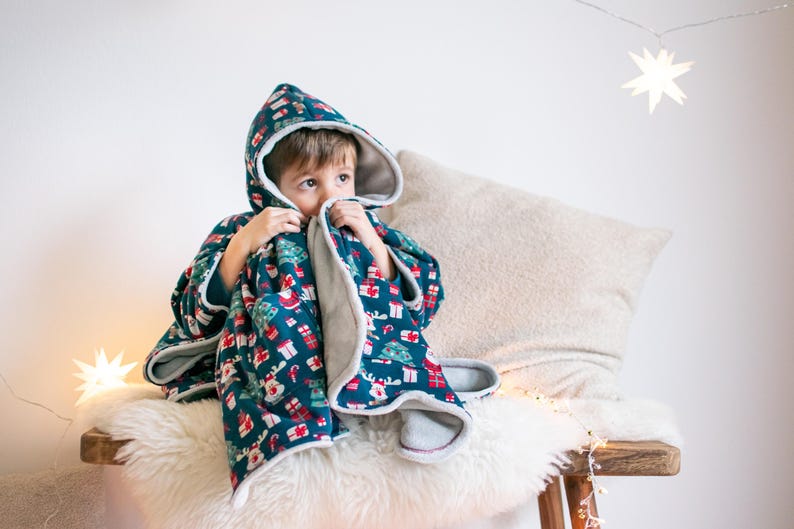 May include: A child wearing a hooded, blue and white fleece blanket with a Christmas print. The blanket has a red, white, and green pattern with images of Christmas trees, presents, and reindeer. The child is sitting on a white furry surface with a beige pillow behind them.