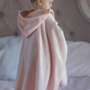May include: A young child wearing a pink hooded blanket with a soft, plush texture. The blanket is draped over the child's shoulders and has a hood that is pulled up over their head.