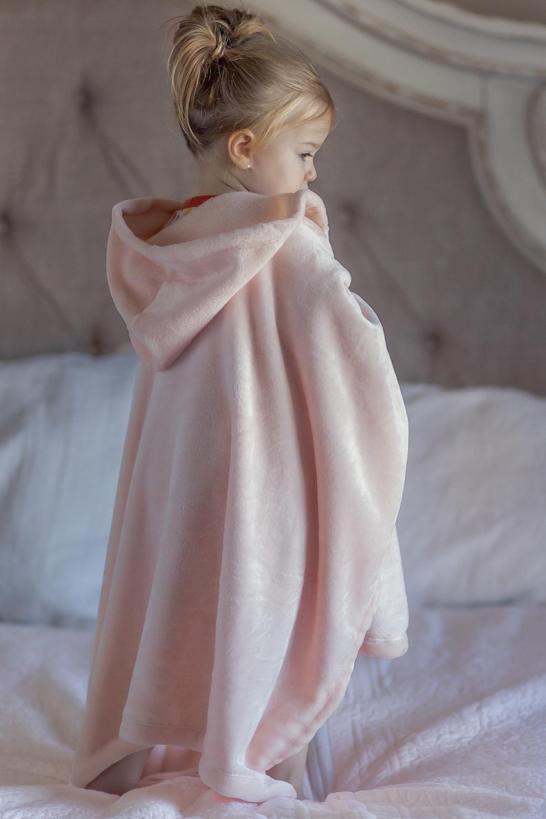 May include: A young child wearing a pink hooded blanket with a soft, plush texture. The blanket is draped over the child's shoulders and has a hood that is pulled up over their head.