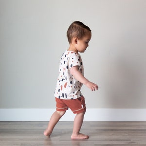 May include: A toddler wearing a white short-sleeved shirt with a brown, black, and white abstract print and brown shorts walks on a wood floor.