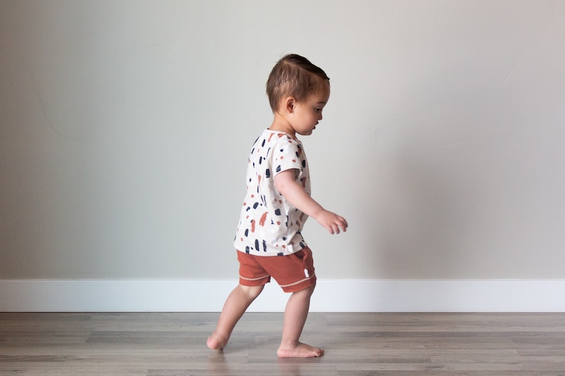 May include: A toddler wearing a white short-sleeved shirt with a brown, black, and white abstract print and brown shorts walks on a wood floor.