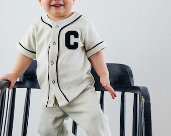 May include: A light gray baseball-style jersey and matching pants set for a baby. The jersey has black trim and a large black "C" on the chest. The baby is standing on a black chair and smiling.