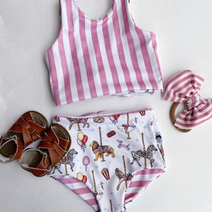 May include: A two-piece swimsuit set with a pink and white striped top and high-waisted bottoms featuring a carousel print. A matching striped bow and brown leather sandals complete the set. The swimsuit is designed for children.