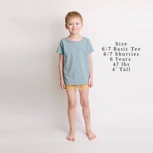 May include: A young boy wearing a light blue t-shirt and tan shorts. The text "Size 6/7 Basic Tee 6/7 Shorties 6 Years 47 lbs 4' Tall" is written in black text on a white background.