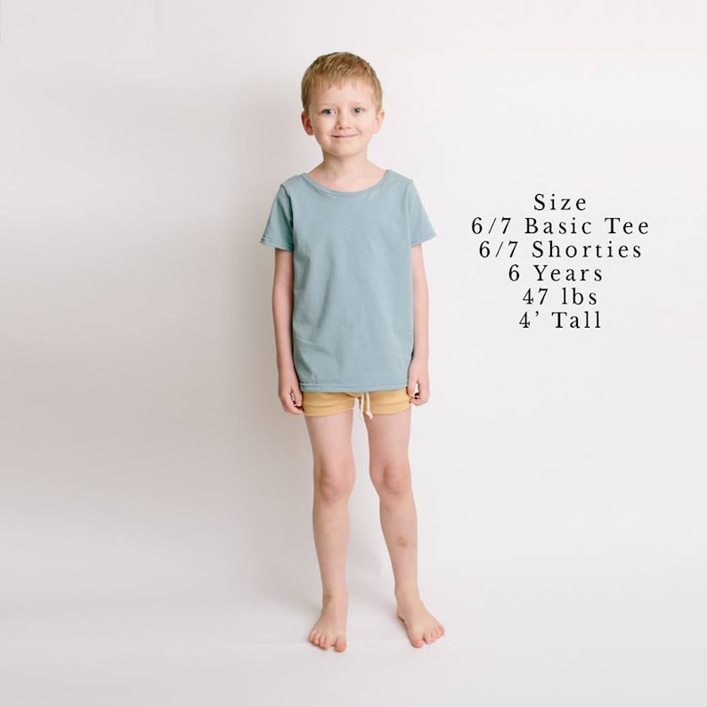 May include: A young boy wearing a light blue t-shirt and tan shorts. The text "Size 6/7 Basic Tee 6/7 Shorties 6 Years 47 lbs 4' Tall" is written in black text on a white background.