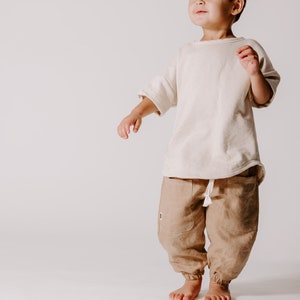 May include: A young child wearing a white t-shirt and brown trousers stands on a white background. The child is looking up and to the right.