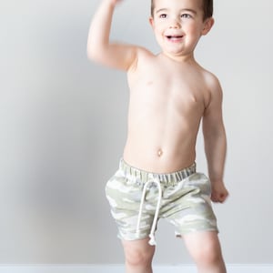 May include: A young boy wearing camouflage print shorts, smiling and raising his right arm in the air.