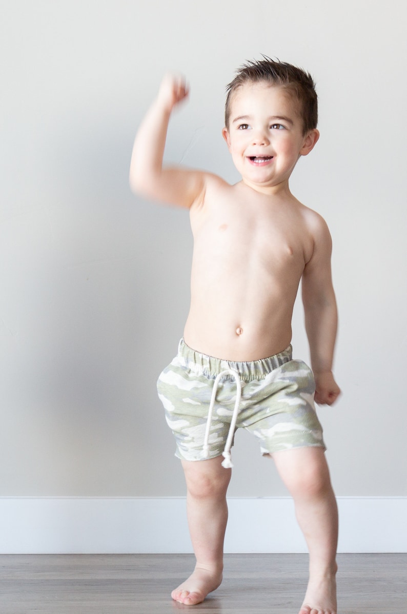 May include: A young boy wearing camouflage print shorts, smiling and raising his right arm in the air.