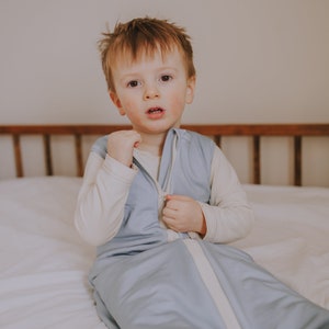 May include: A young child wearing a light blue sleep sack with a white zipper. The sleep sack has a white trim around the zipper and the neckline. The child is sitting on a white bed with a wooden headboard.