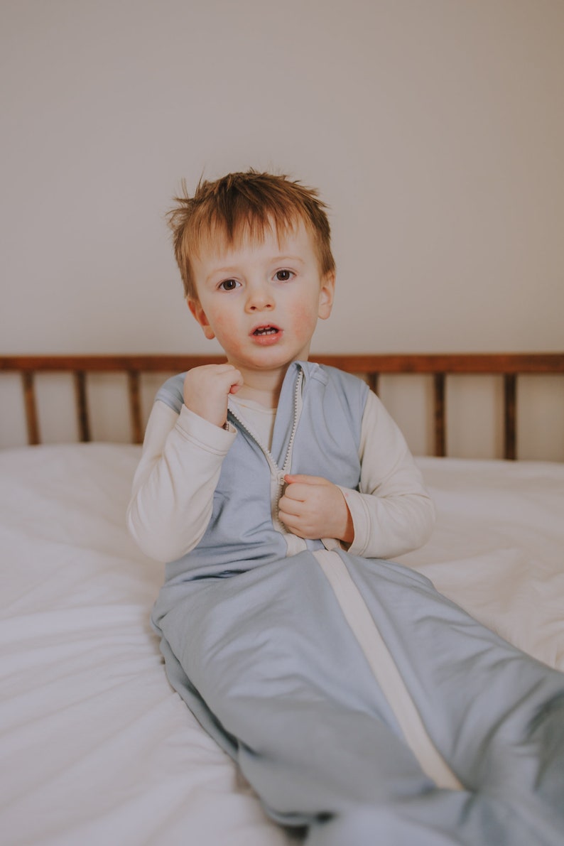 May include: A young child wearing a light blue sleep sack with a white zipper. The sleep sack has a white trim around the zipper and the neckline. The child is sitting on a white bed with a wooden headboard.