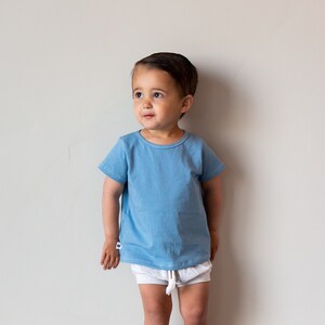 May include: A young child wearing a light blue short-sleeved t-shirt and white shorts stands in front of a white wall.